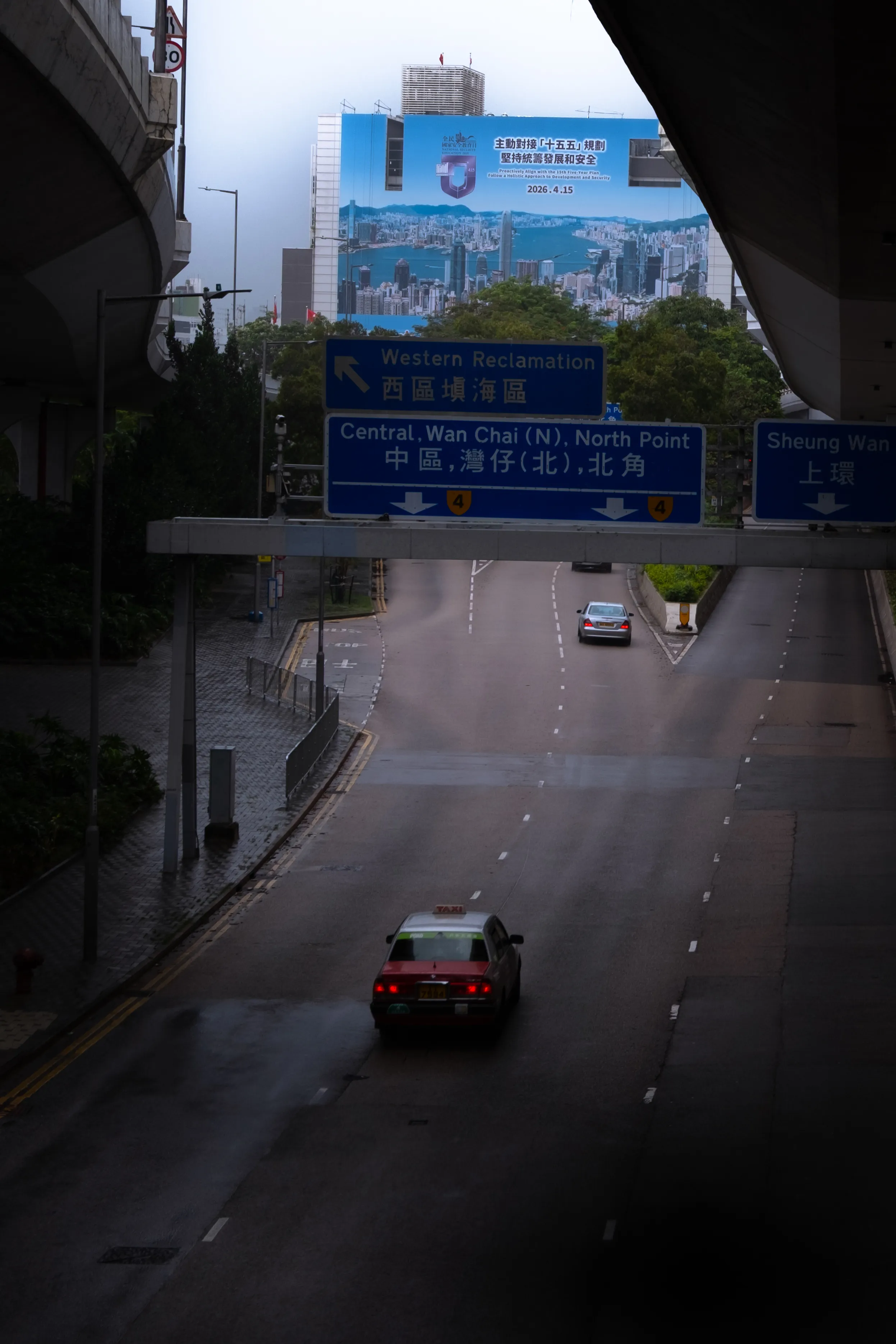 A red Hong Kong taxi on an empty multi-lane road beneath flyovers, with blue direction signs for Western Reclamation, Central and Sheung Wan, and a large government billboard above