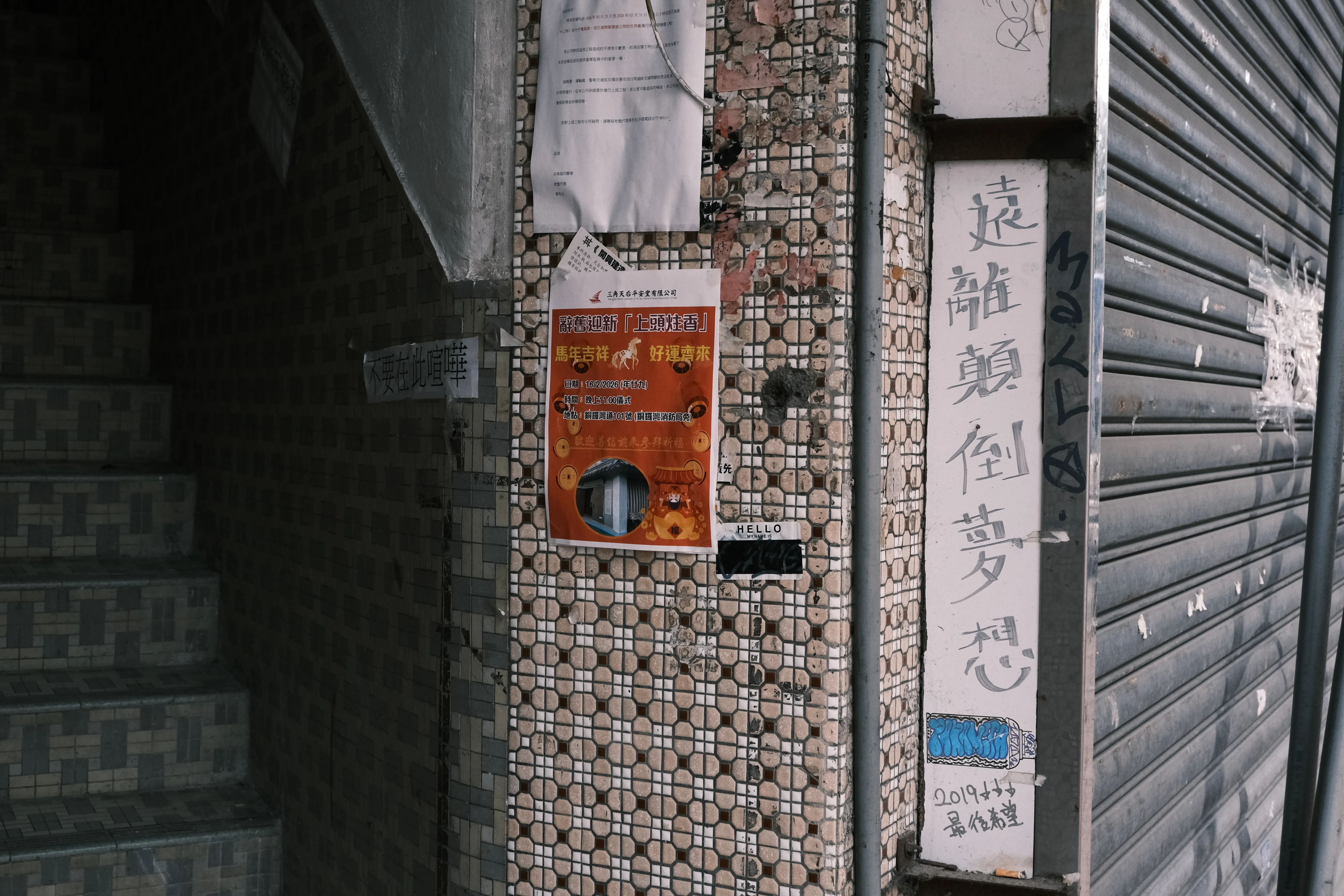 A mosaic-tiled stairwell entrance beside a metal shutter, with a flyer on the wall and Chinese characters handwritten on a white pillar