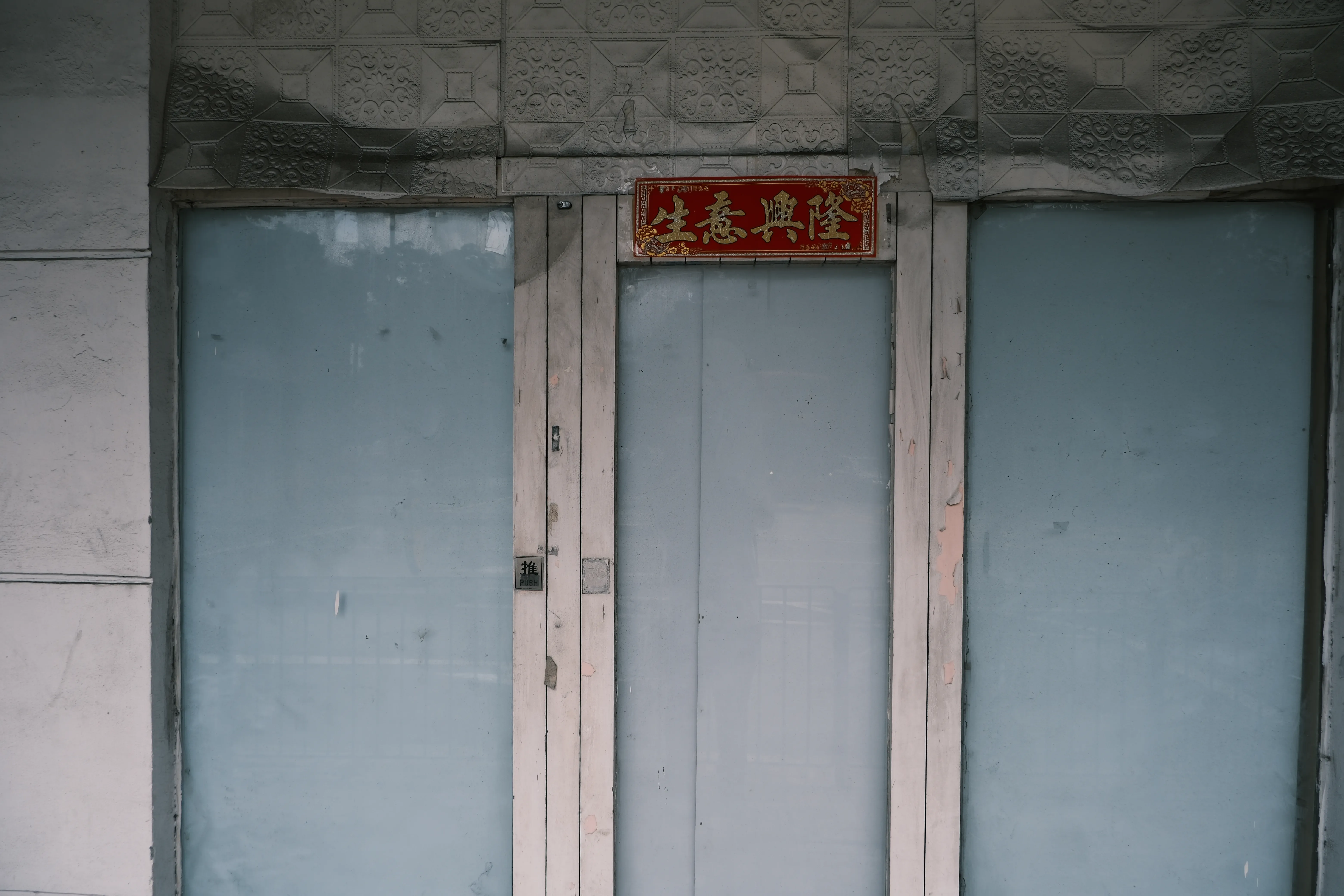 A closed shopfront of pale blue panels under grey pressed-metal tiles, with a small red plaque reading 生意興隆