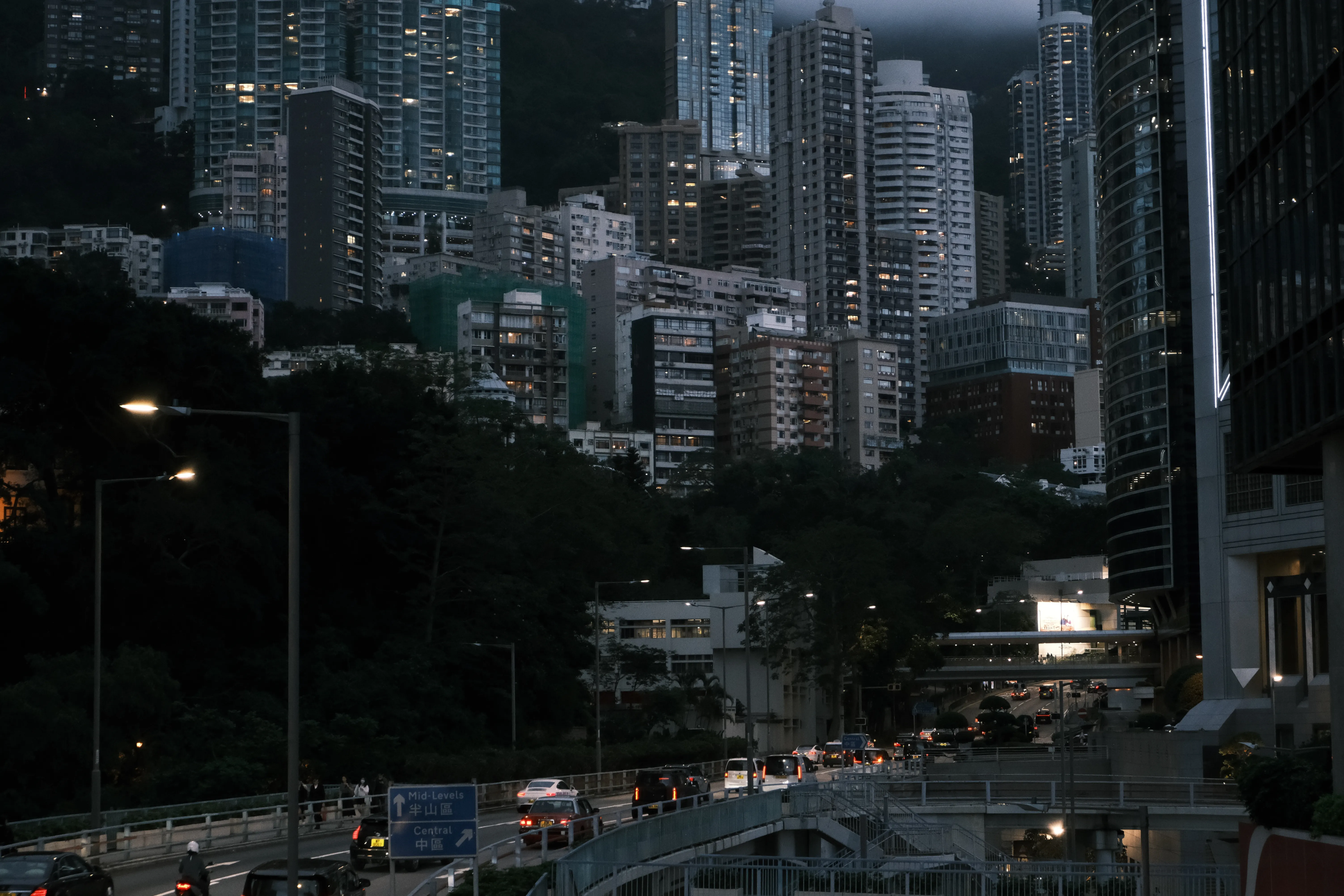 Dense high-rise towers climb a dark, forested hillside at dusk above a busy road with a sign pointing to Mid-Levels and Central