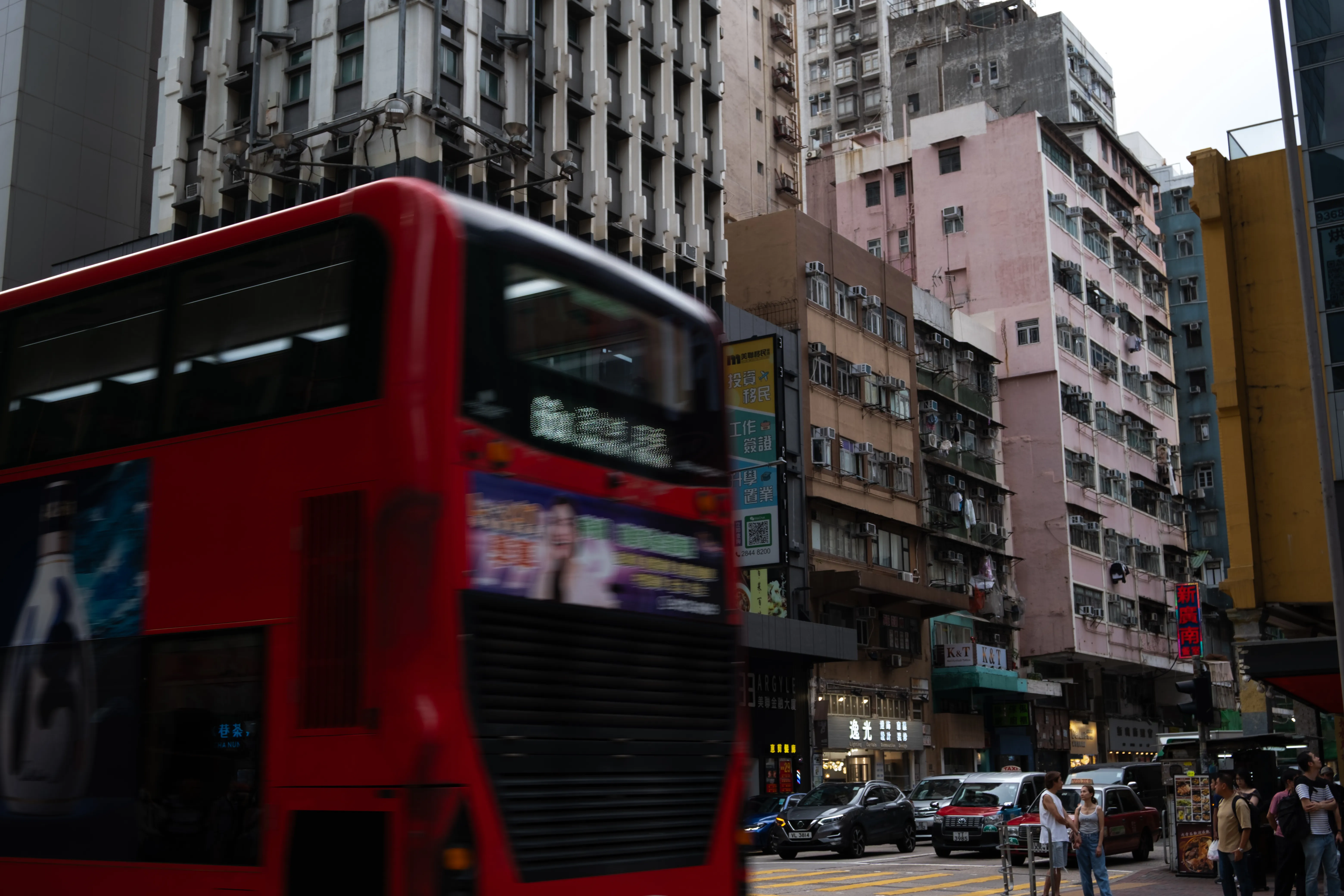 A red double-decker bus blurs past a street corner lined with pink and beige tenement buildings covered in air-conditioning units