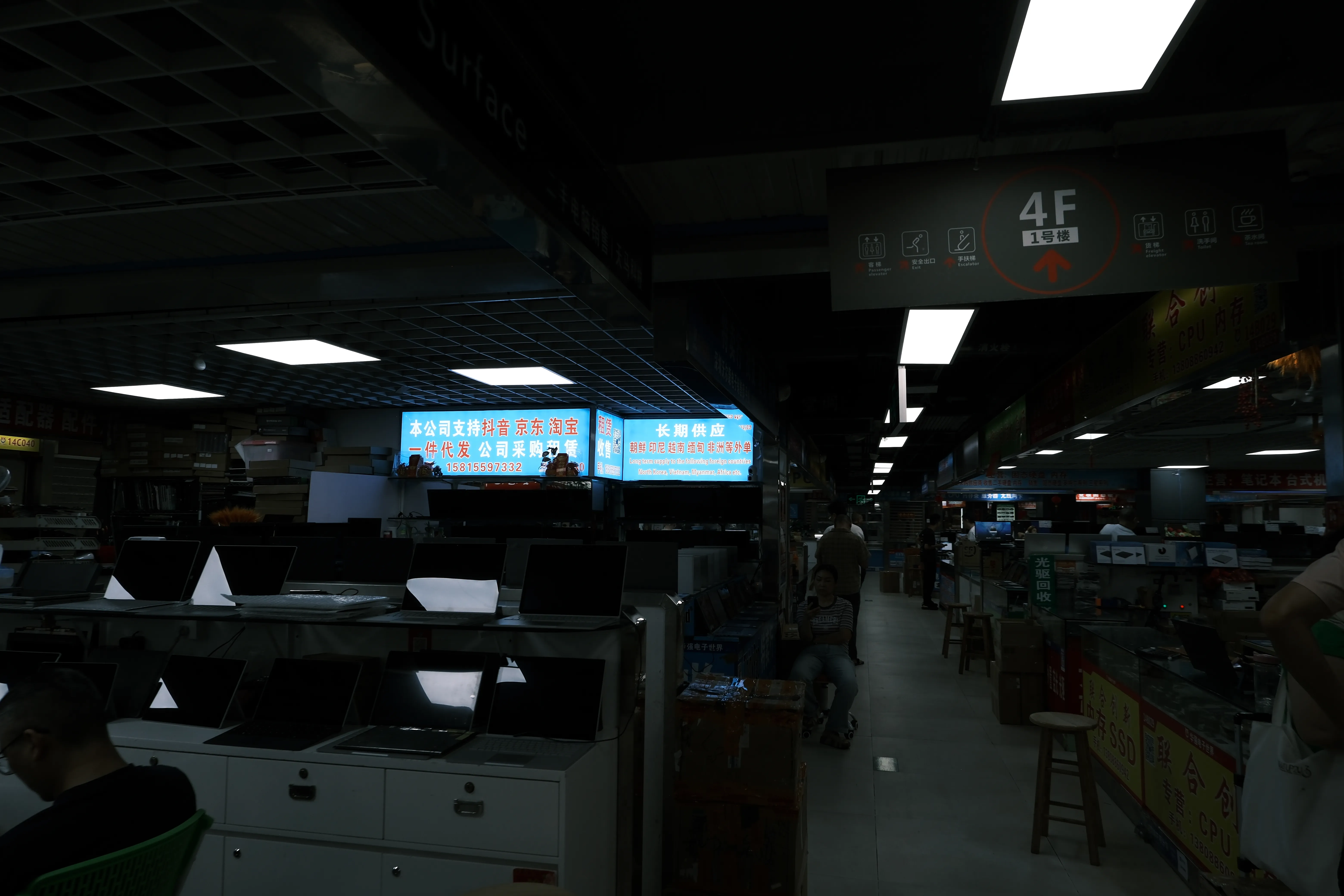 A dim fourth-floor electronics market hall with rows of laptops on display counters, bright advertising screens and a few vendors