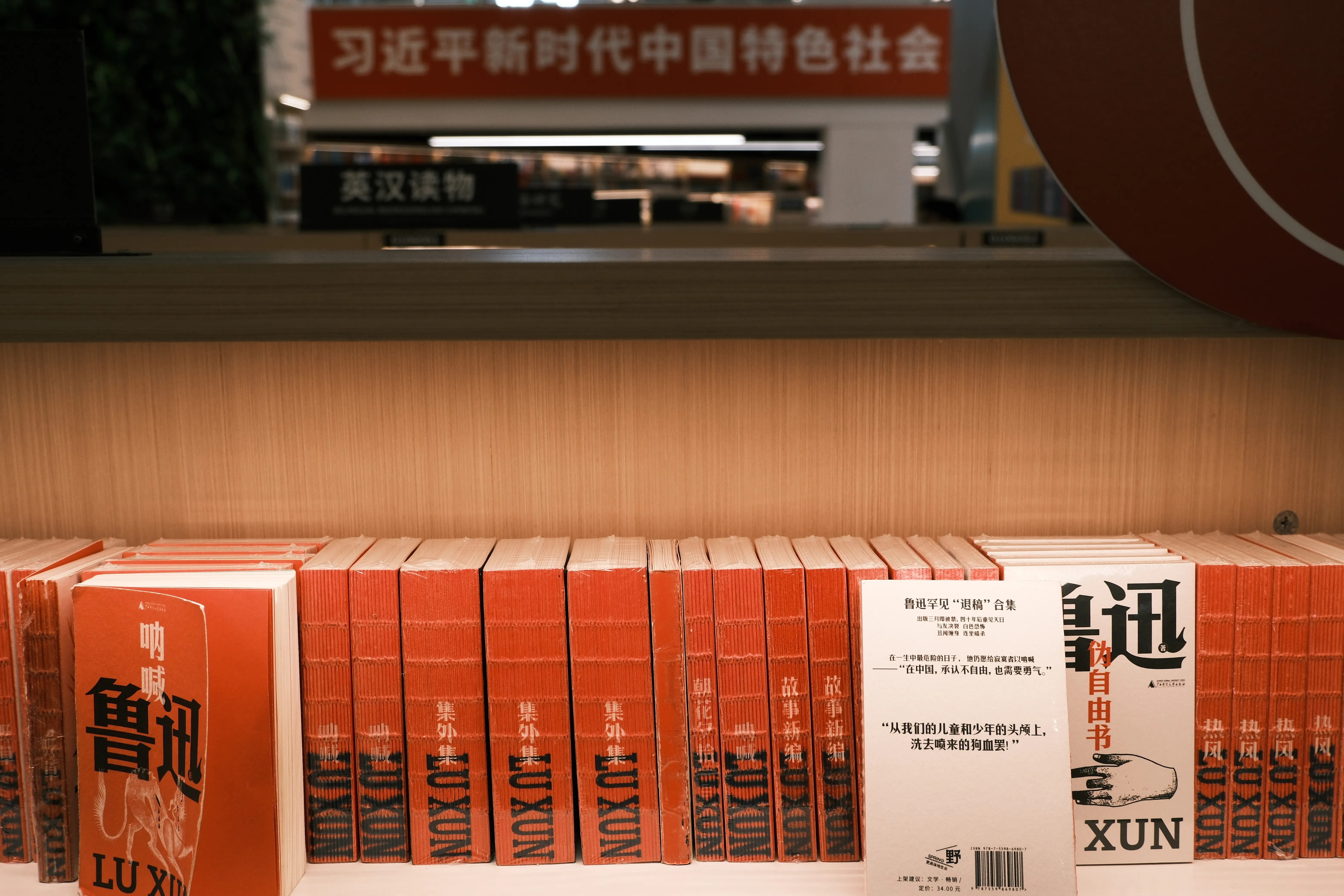 A shelf of orange Lu Xun paperbacks in a bookstore, with a red banner about Xi Jinping Thought visible in the background