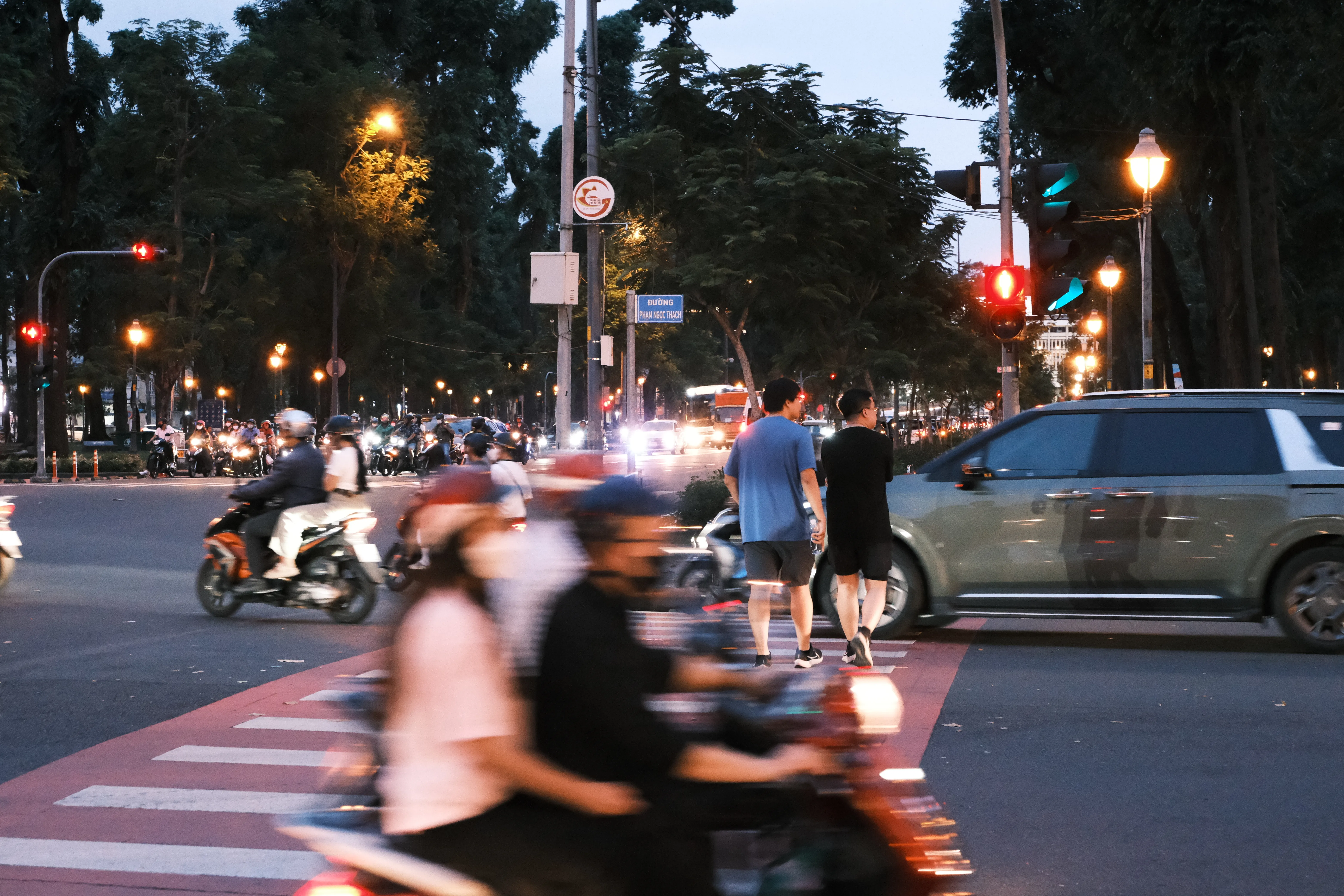 Motorbike riders blur through a crosswalk at dusk on a tree-lined boulevard, as two men cross behind them and a car waits at a red light